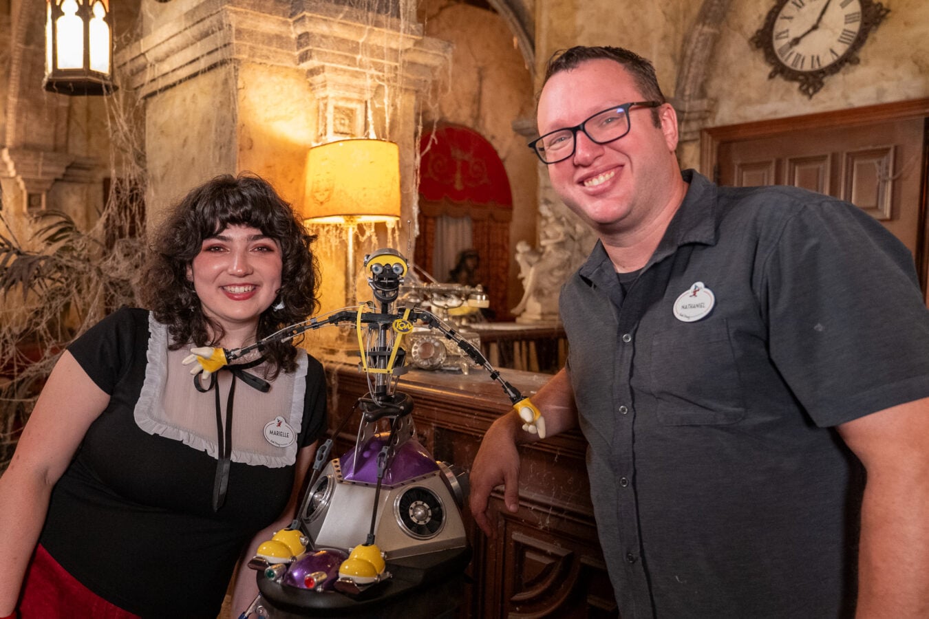Two people smile beside a small robot model on a table in a Halloween themed room with cobwebs and antique decor behind them