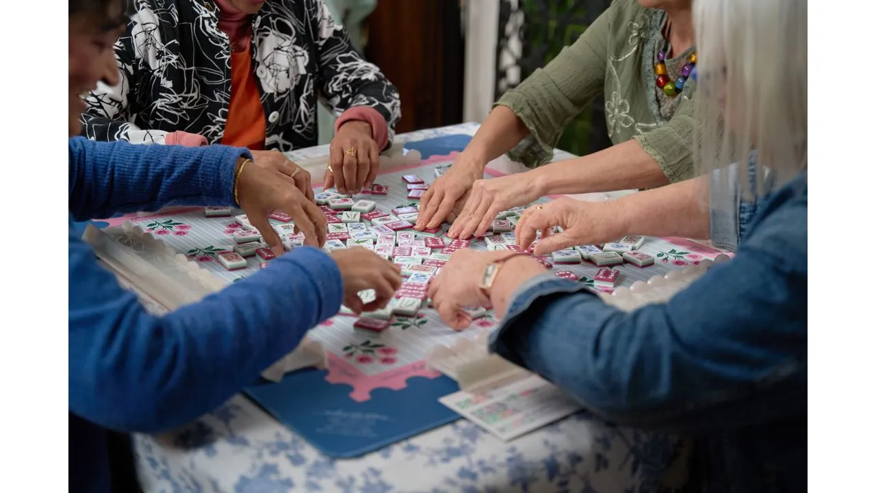 Group of people gathered around a table playing a tile based game laying out small square tiles with pink backs