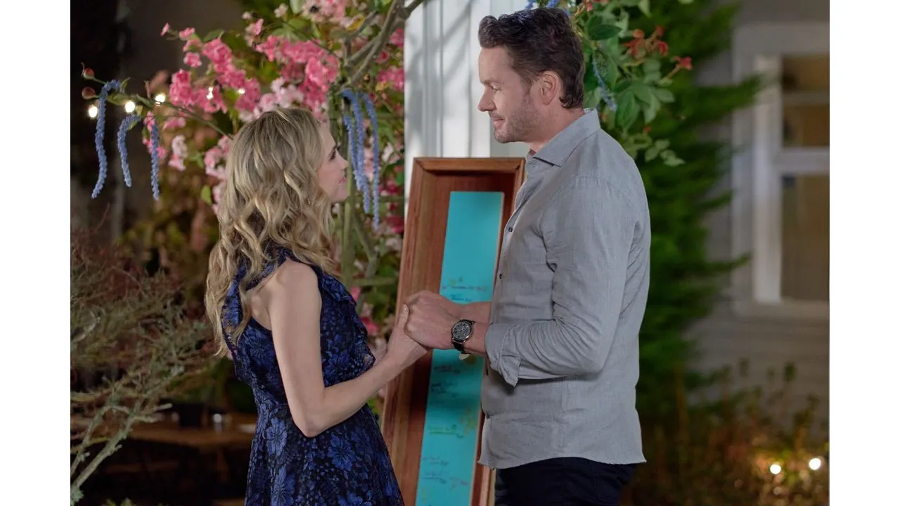 Couple facing each other holding hands in a garden with pink flowers and a teal display board in the background