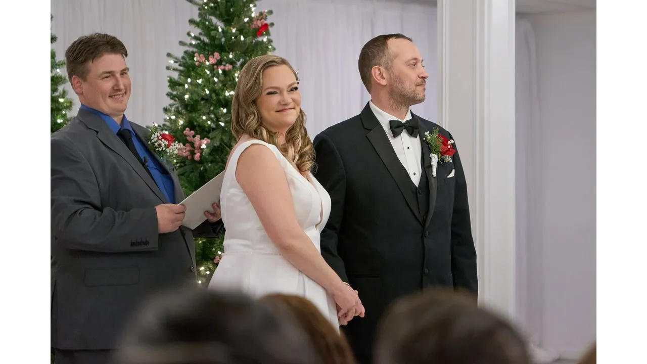 Bride in white gown and groom in a tuxedo hold hands during their ceremony officiant smiling in the background beside Christmas trees in a festive venue