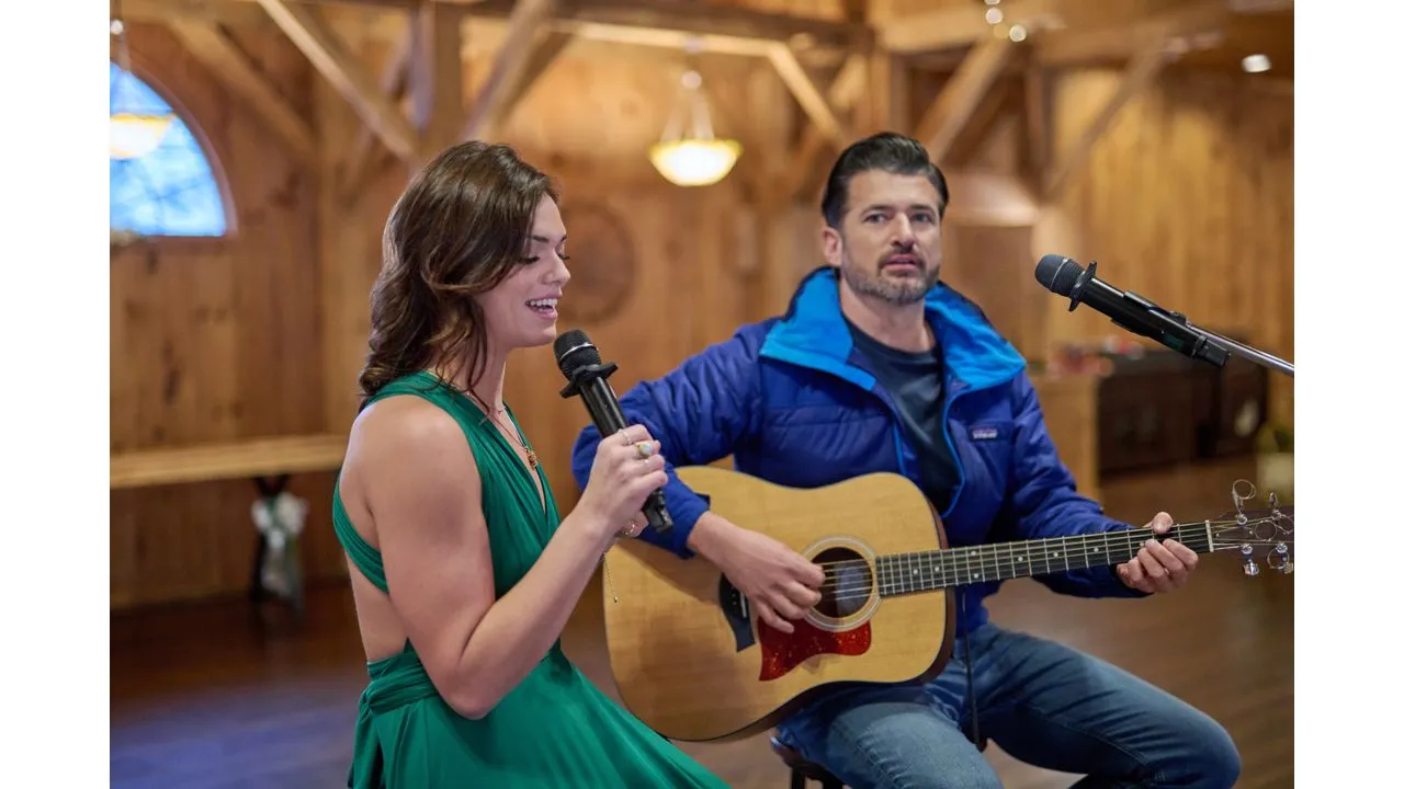 A woman in a green dress sings into a microphone while a man in a blue jacket plays an acoustic guitar beside her in a cozy wooden venue