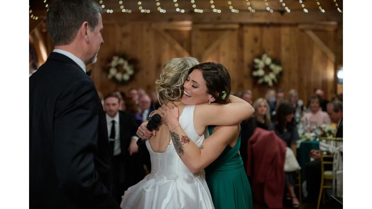 Bride in a white gown embraces a guest in a green dress at a wedding reception smiling warmly as others watch in a rustic venue with string lights on the ceiling