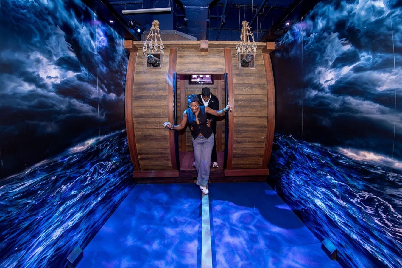Person in a blue vest and jeans exits a wooden barrel shaped ride capsule with a helper behind set against a wall mural of turbulent ocean waves