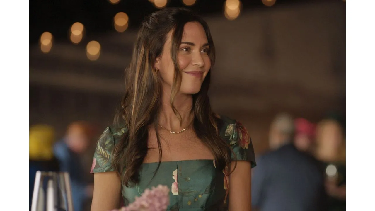 Woman in a green floral off shoulder dress smiling at a social event with warm bokeh lights in the background