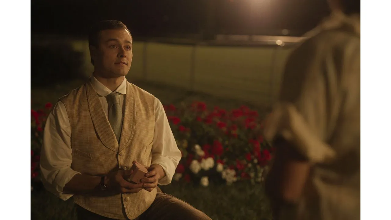 Man in a beige vest and tie sits outdoors at night holding a small box and talking to a blurred person with red flowers in the background