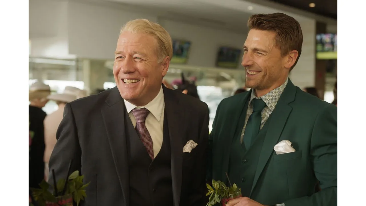 Two men in suits smiling and chatting at a social event one in a black suit with burgundy tie the other in a green suit