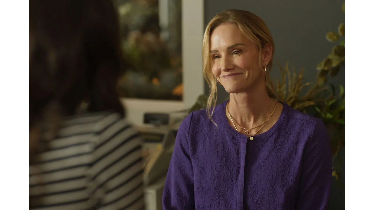 Smiling blonde woman in a purple textured jacket talks with someone indoors