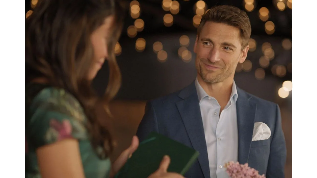 Man in a blue suit smiles at a woman holding a green booklet during a festive evening with warm bokeh lights in the background