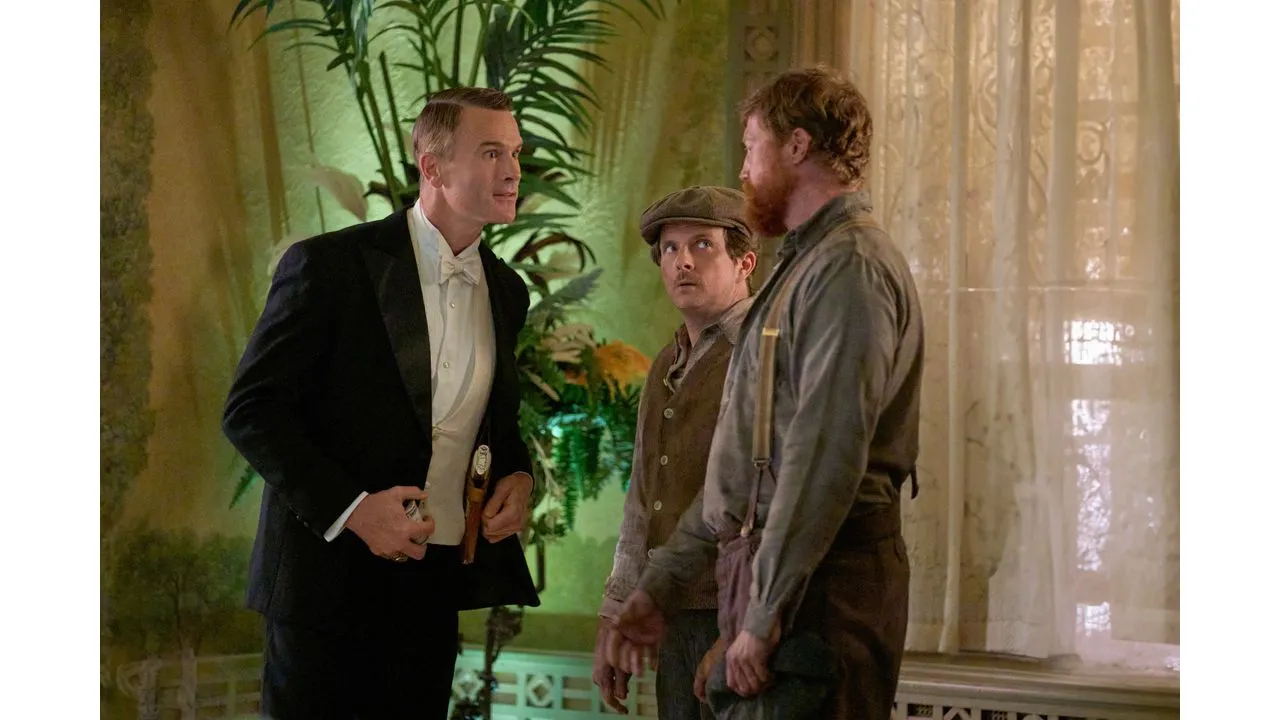 Three men in an opulent plant filled room converse a tuxedoed man on the left and two workers in caps and suspenders on the right