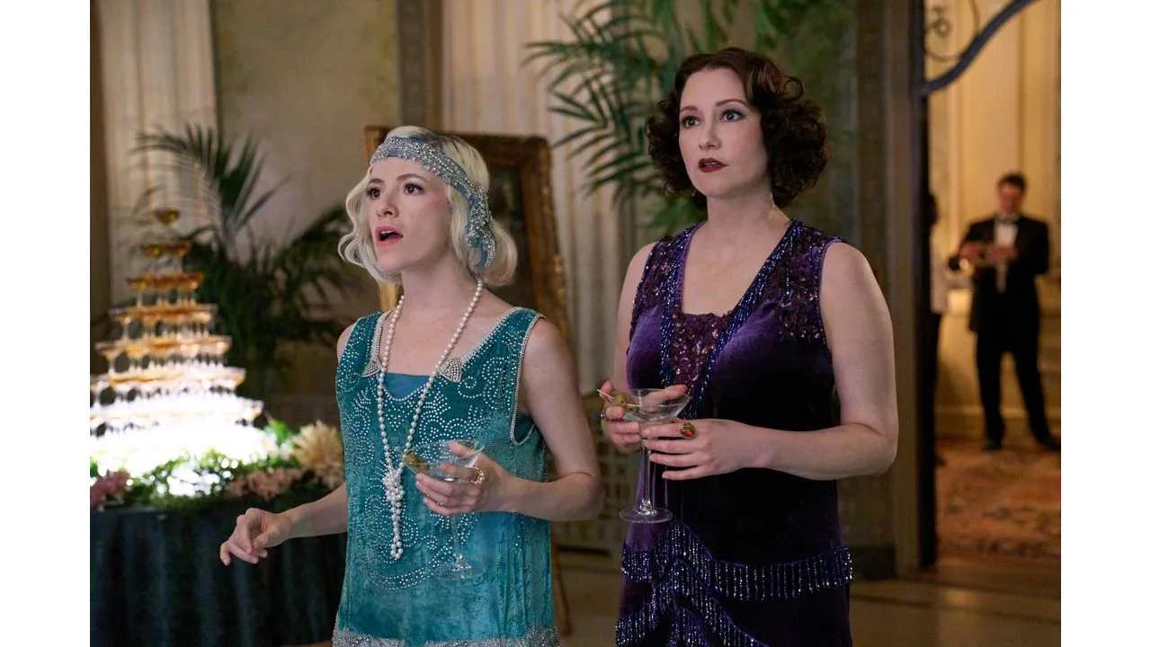 Two women in 1920s flapper dresses hold champagne glasses at an elegant party by a dessert table in the background
