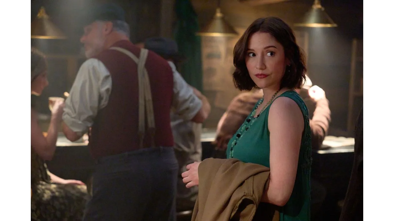 Woman in a teal dress turns to look at the camera while holding a tan coat in a dimly lit bar with patrons in the background