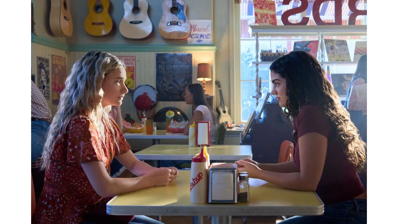 Two young women sit across from each other at a retro diner table chatting with condiments between them under guitar decor on the wall