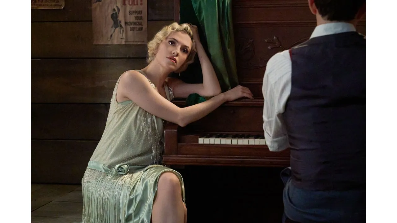 Blonde woman in a pale green fringe dress leans on an upright piano watching a man in a vest from the side