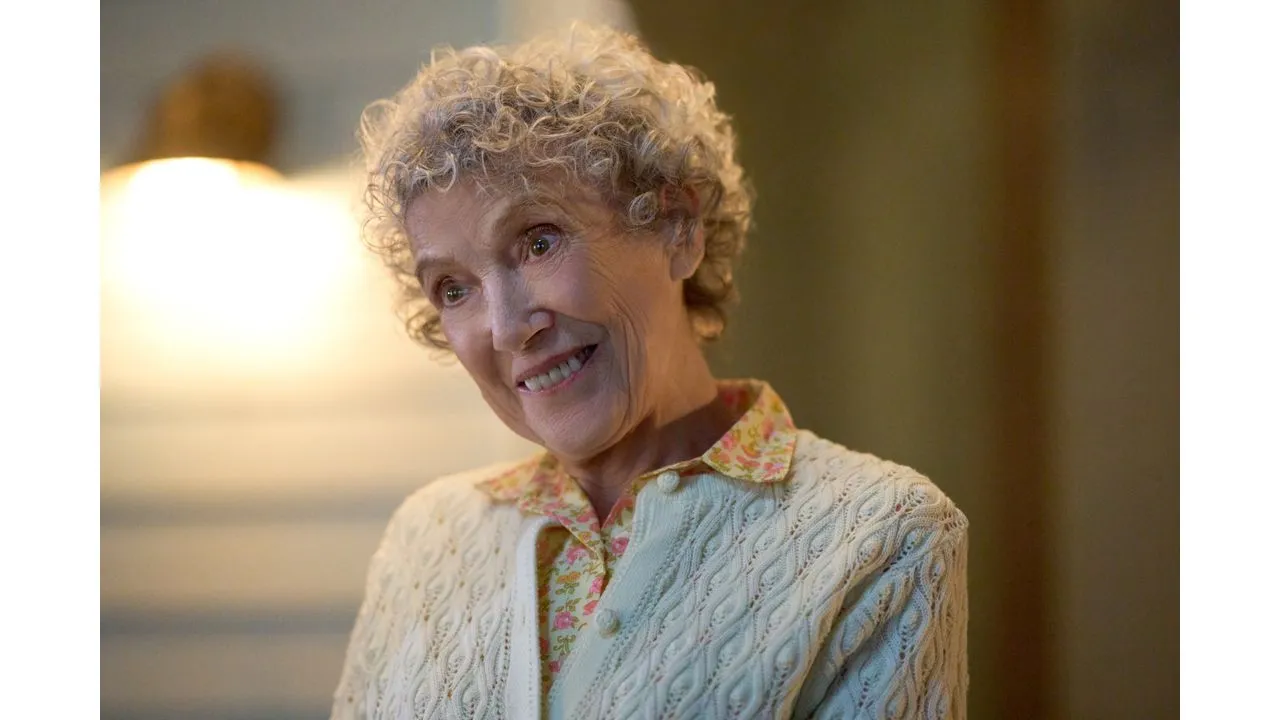 Smiling elderly woman with curly gray hair wearing a cream knit cardigan over a floral blouse in warm indoor light