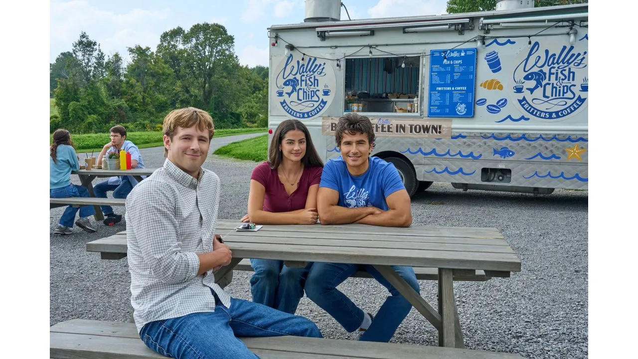 Three friends sit at a wooden picnic table in a gravel lot posing for the camera with a fish and chips truck in the background