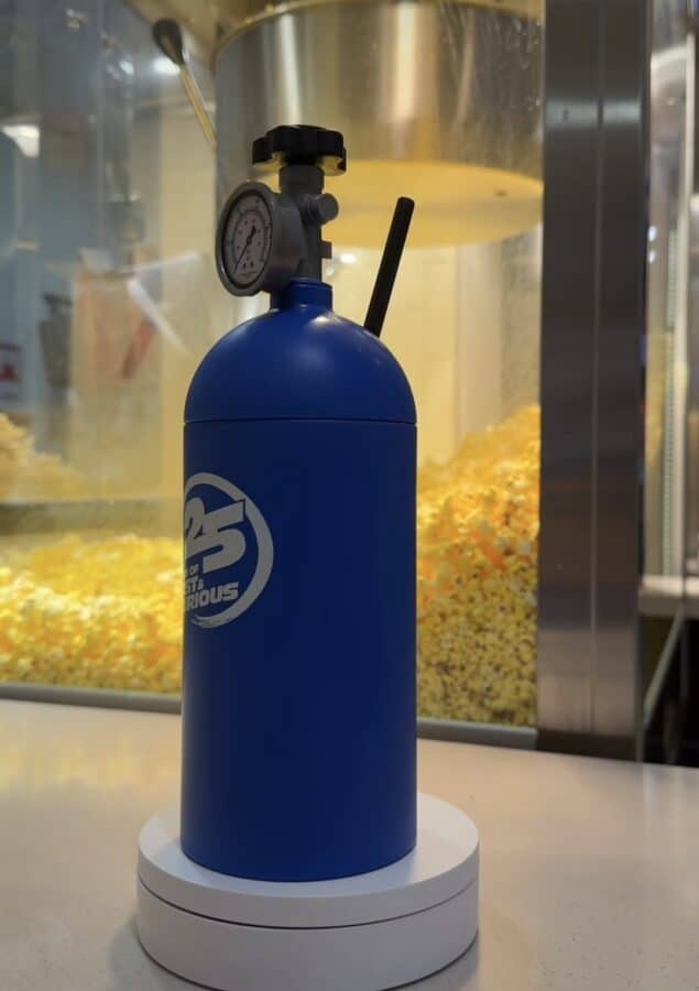Blue pressurized gas cylinder with a gauge and valve on top placed on a white pedestal against a popcorn machine backdrop