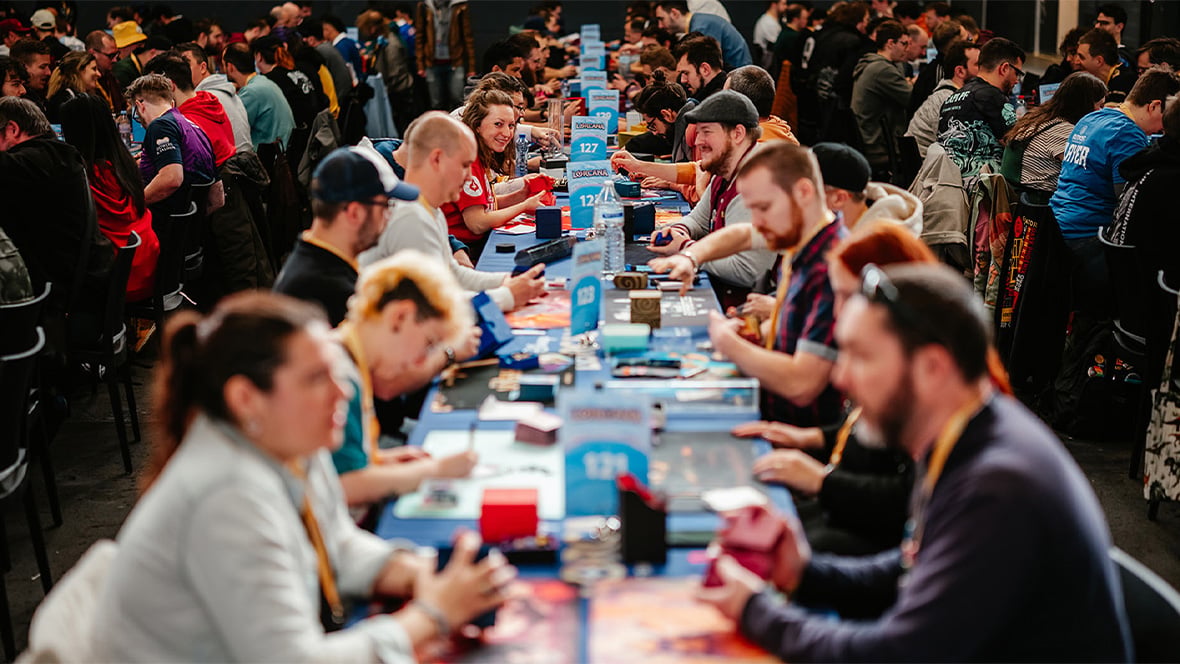 Long row of players seated opposite each other at a gaming tournament table concentrating on cards with a cheering crowd in the background