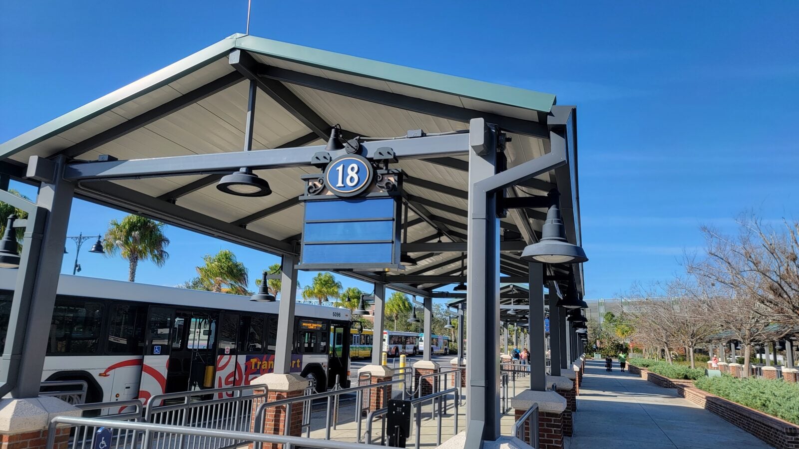 Bus stop shelter with a blue numbered sign reading 18 and a parked bus nearby under a clear blue sky