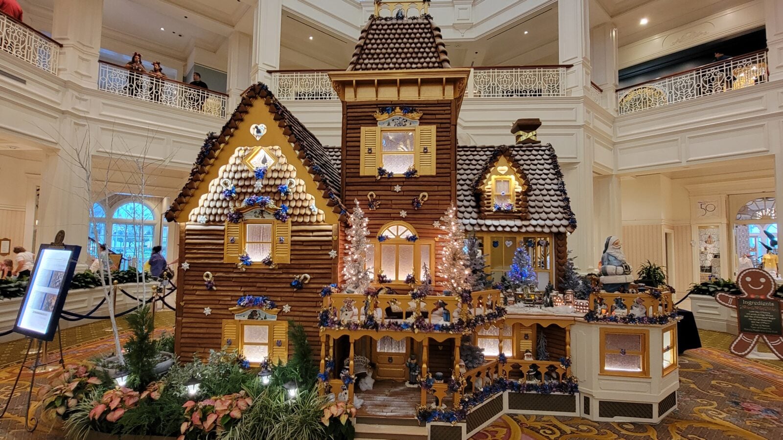 Large festive gingerbread house display in a bright hotel atrium with decorative blue and white accents and surrounding walkway railings versus balconies above