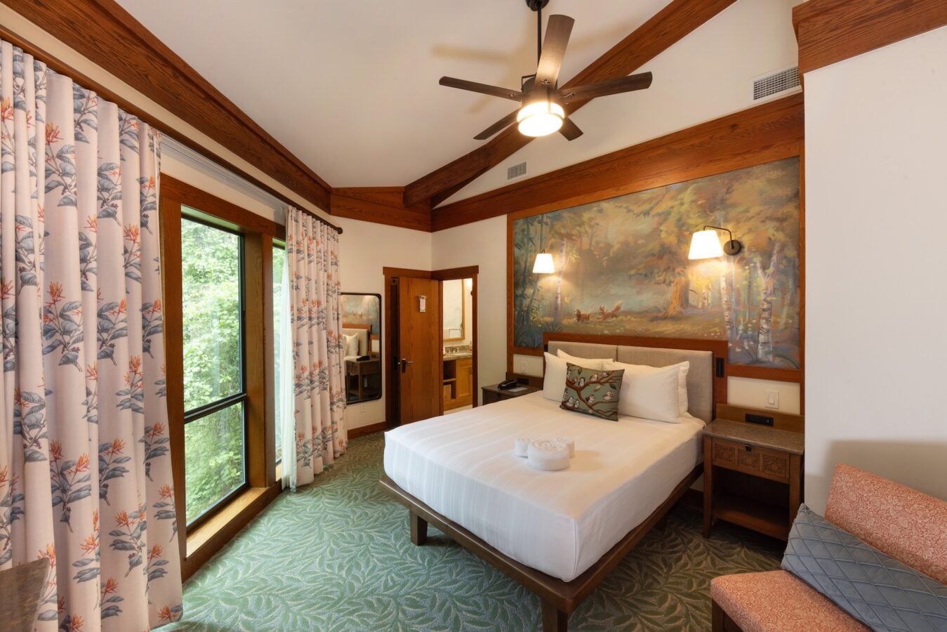 Cozy hotel bedroom with a king bed forest themed painting above wood beams ceiling fan and large windows with floral curtains