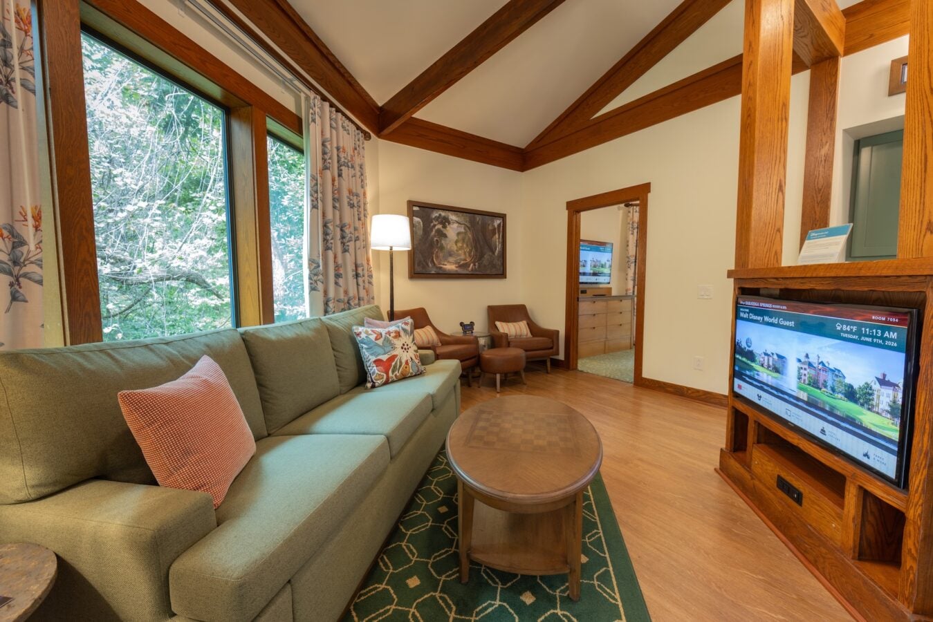 Cozy living room with a green sectional floral pillows tall windows showing trees and exposed wooden beams