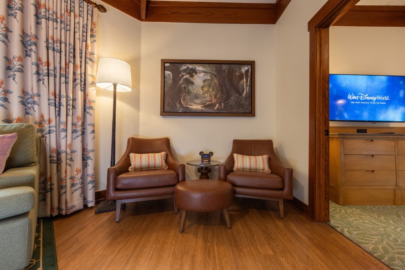 Cozy hotel seating area with two brown leather chairs striped pillows and a round ottoman around a small side table under a wall painting of a forest