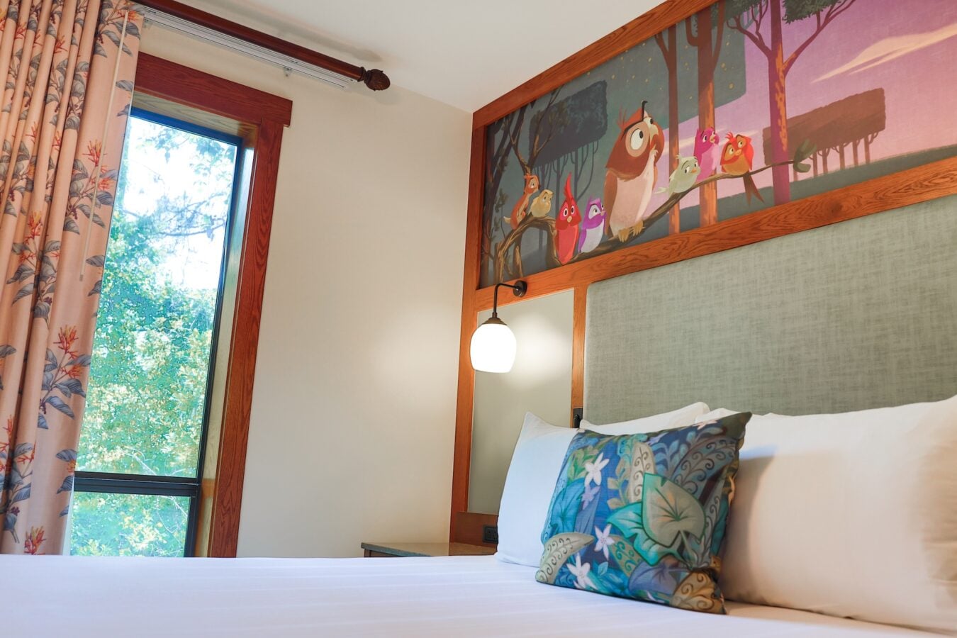 Bed with white sheets and colorful patterned pillows under a wooden headboard topped by a mural of cartoon birds on a branch window with pink floral curtains to the left