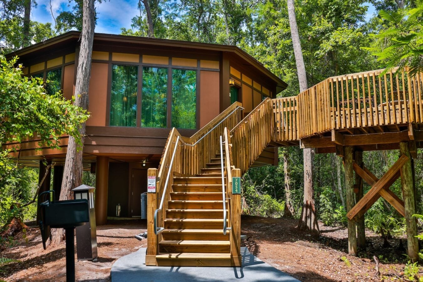 Wooden treehouse on stilts with large glass windows and a wraparound deck reached by a central wooden staircase among tall trees in a forest