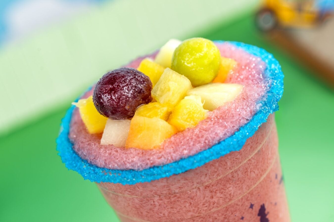 2026WDWCoolKidsSummerTreats31620x1080 Fantasy Land News Pink fruit smoothie in a glass with a blue sugar rim topped with pineapple melon grapes and other diced fruits