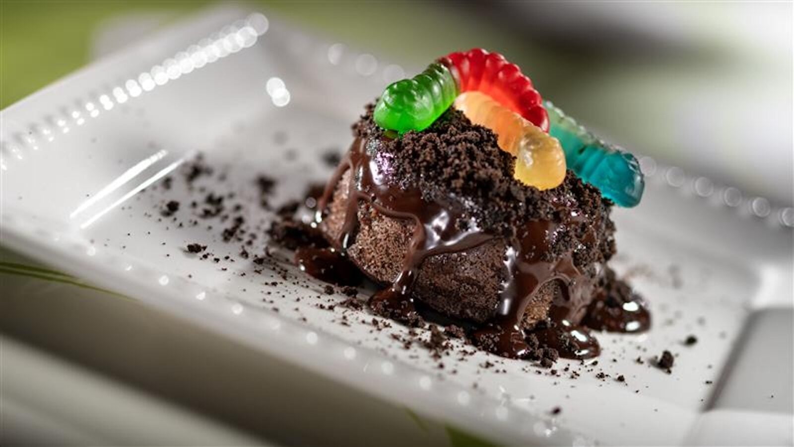 2026WDWCoolKidsSummerTreats6 Fantasy Land News Chocolate dessert on a white plate topped with colorful gummy worms and crumbled cookies with melted chocolate drizzle