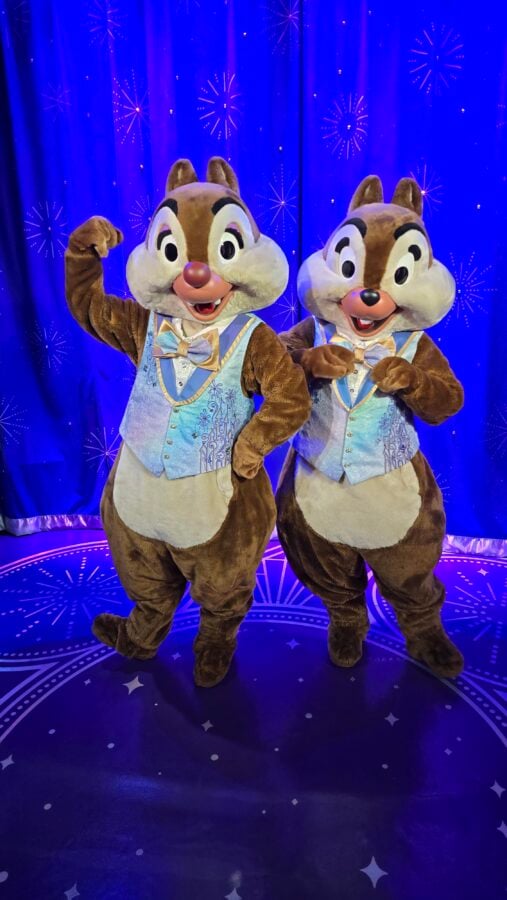 20260501095202 Fantasy Land News Two chipmunk mascots in matching blue vests pose and wave on a blue stage with a fireworks backdrop