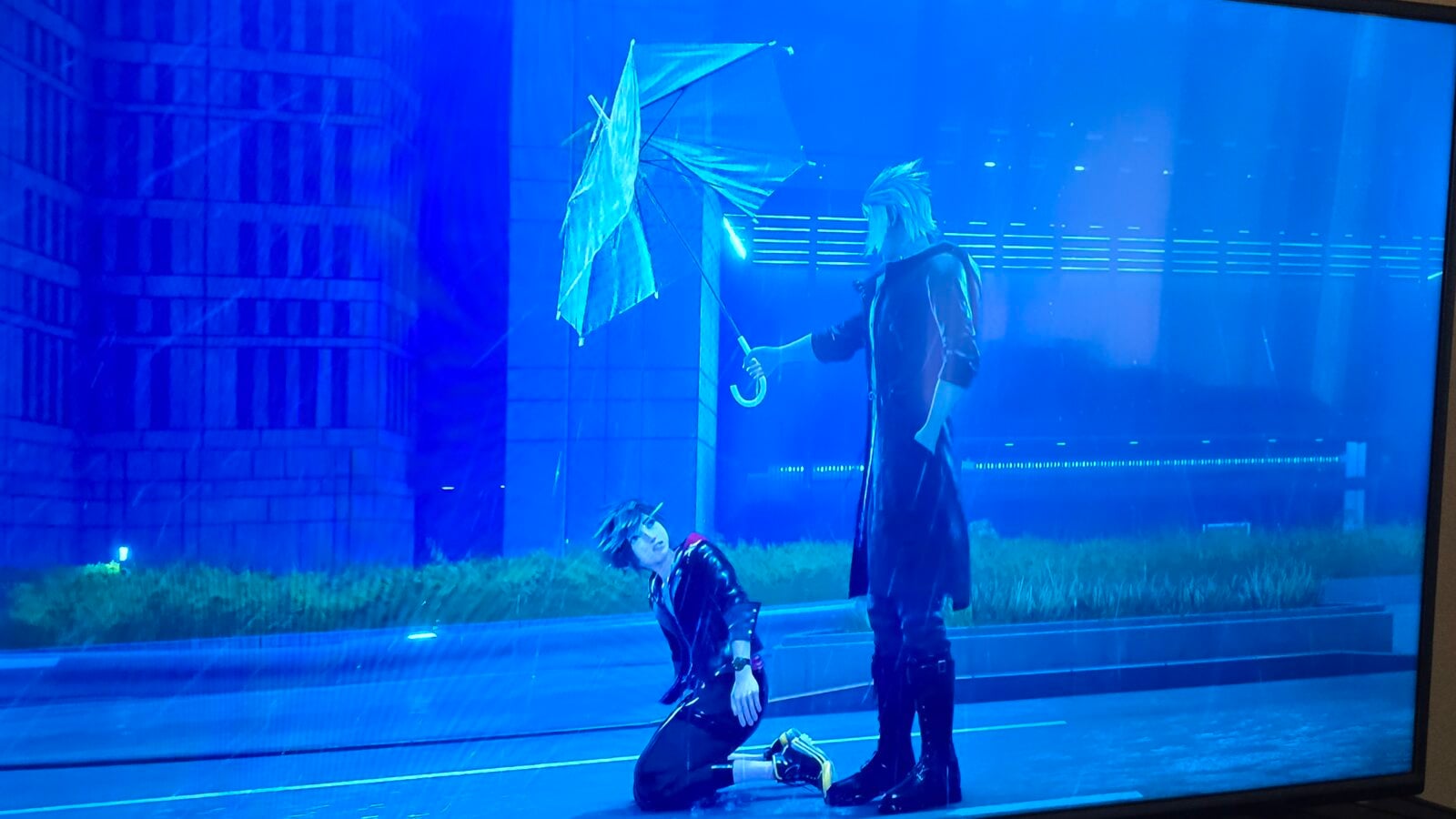 Cosplayers on a blue lit stage one kneels in black outfit holding a translucent umbrella another stands beside wearing gloves and a dark coat
