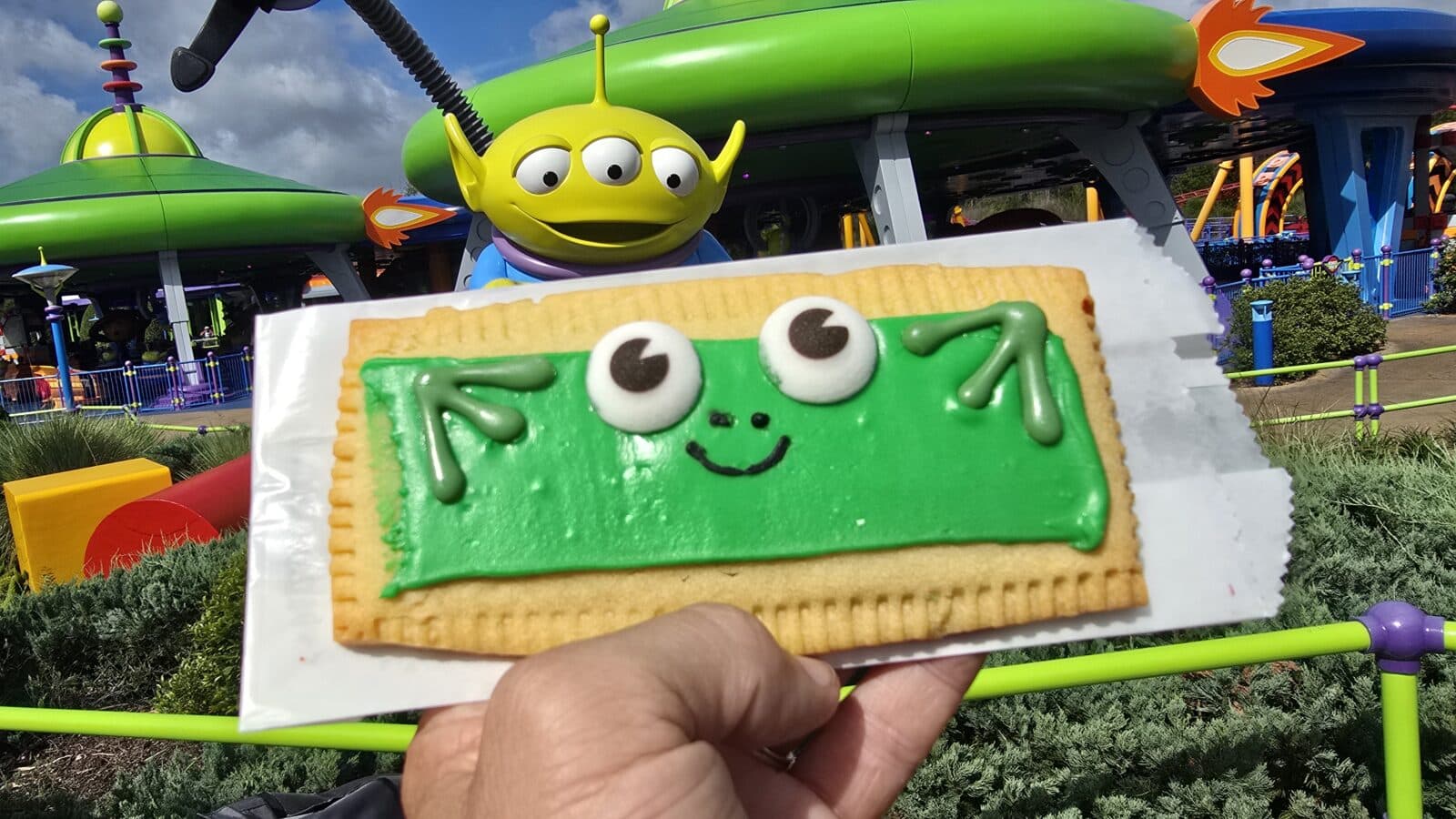 A green frog faced cookie held up in the foreground with a colorful theme park ride and a large yellow alien figure in the background