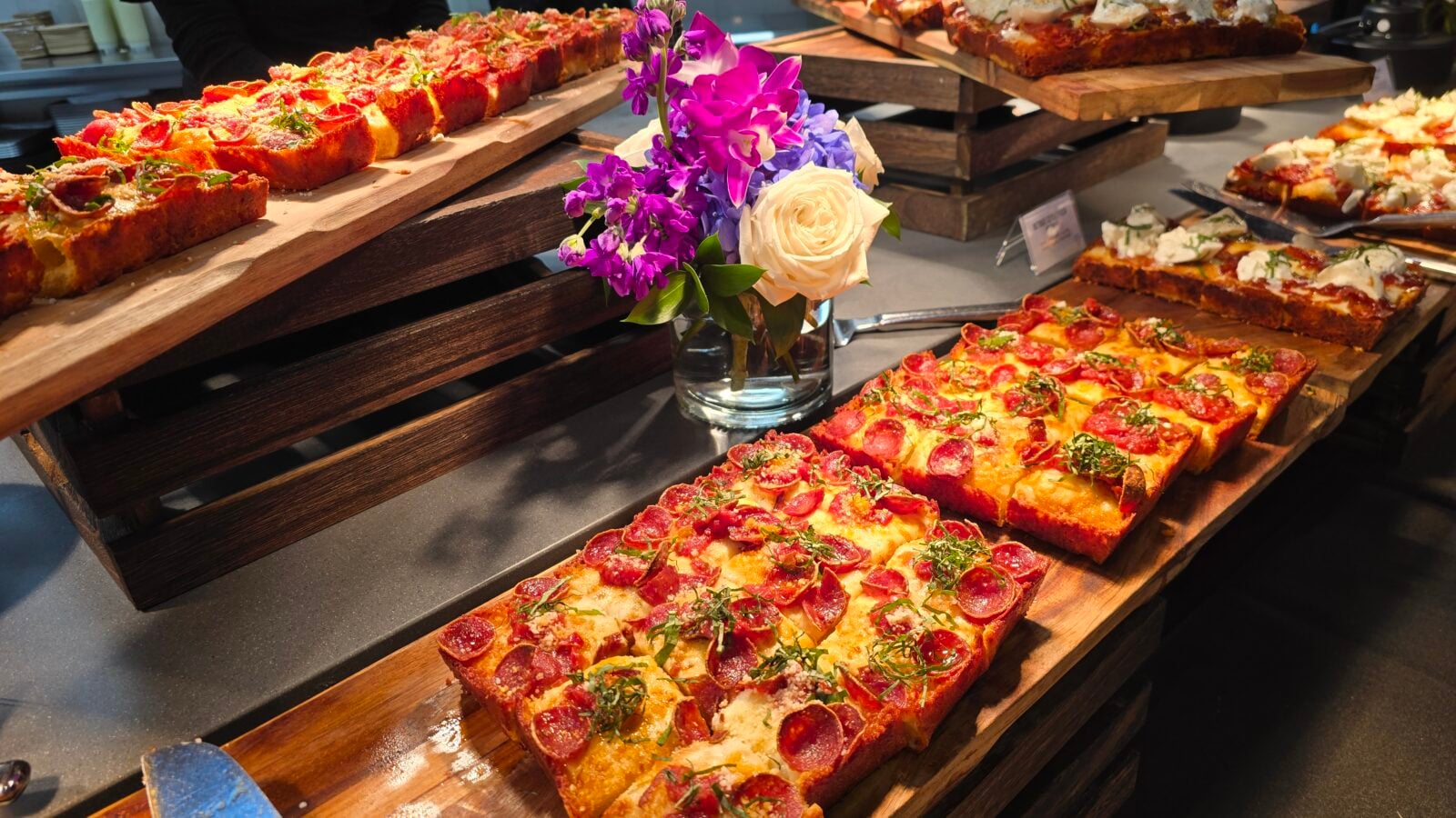 Rows of square pepperoni pizza slices on wooden boards with a bouquet of purple and cream flowers in the center in a buffet setup