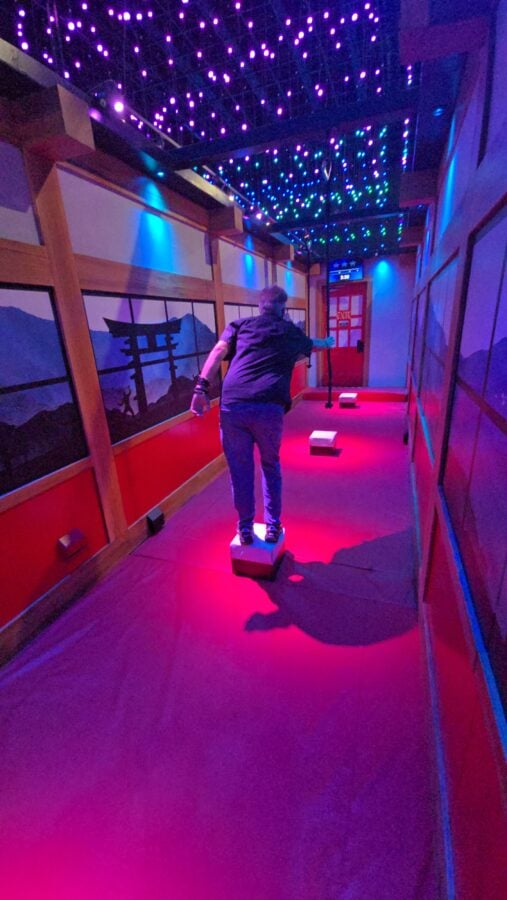 Person standing on small white stepping blocks in a dimly lit hallway with colorful purple and blue ceiling lights giving an immersive glow lit exhibit vibe