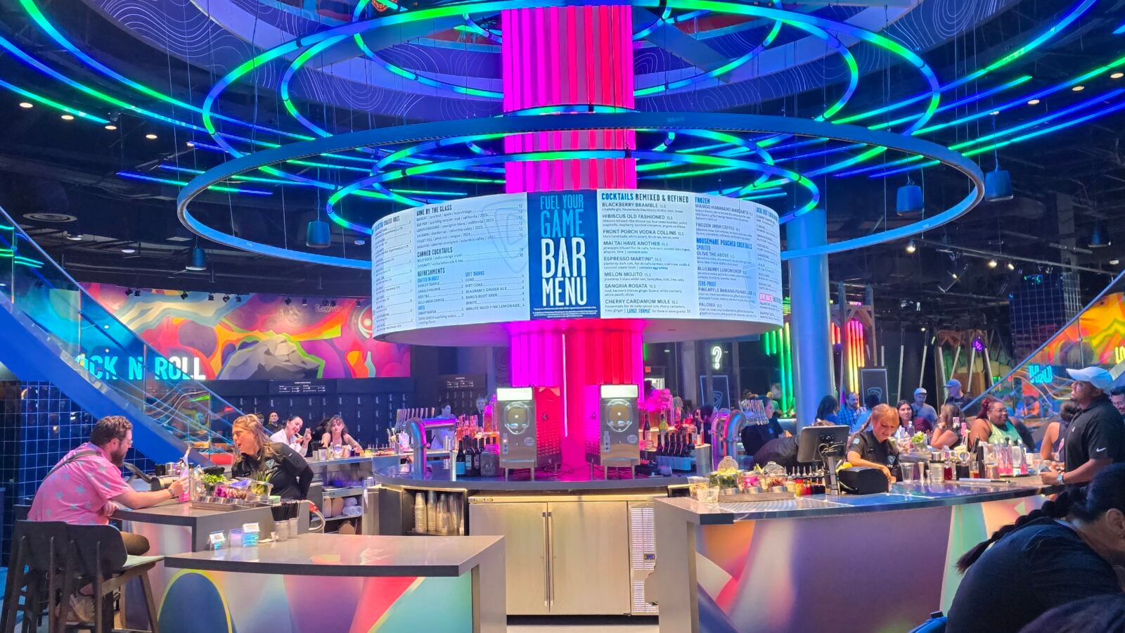 Vibrant bar interior with neon neon rings overhead a central circular menu display and customers seated at the counter enjoying drinks and food
