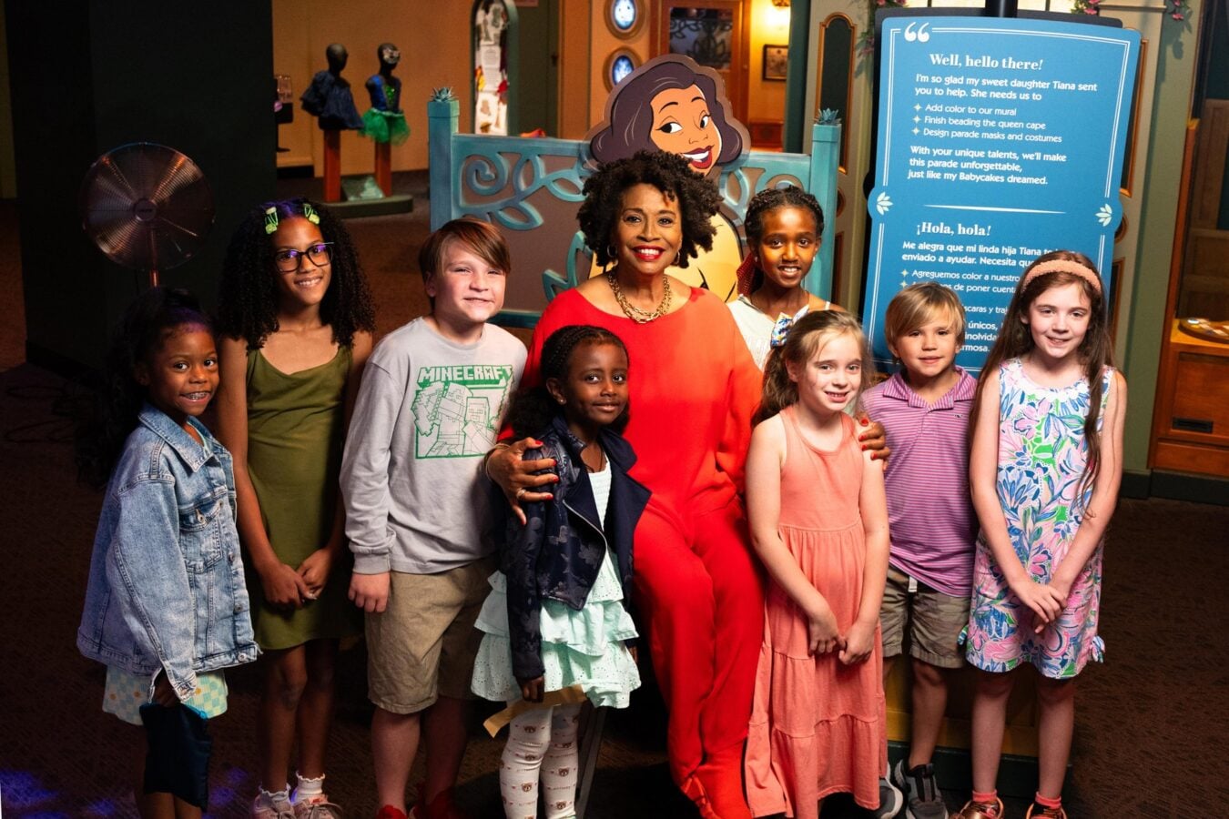 260605TCMIJeniferLewisTiana30 Fantasy Land News Smiling woman in red surrounded by children posing for a group photo in a museum exhibit