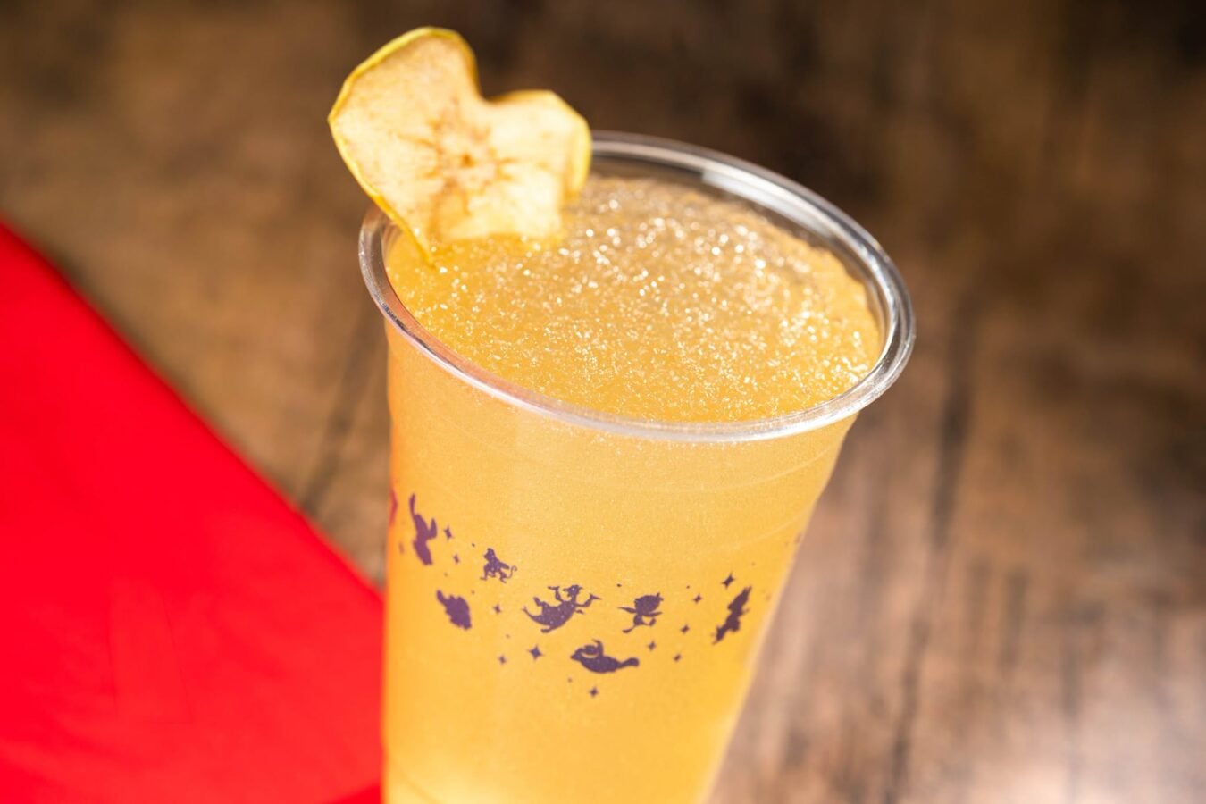71821294910301330761914597406163960879373457n Fantasy Land News Tall clear cup of orange juice or smoothie with a dried apple slice garnish on a wooden table red fabric in the corner