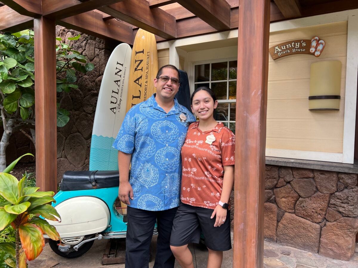 Two people smile for a photo in Hawaiian shirts standing beside surfboards at a beach house entrance
