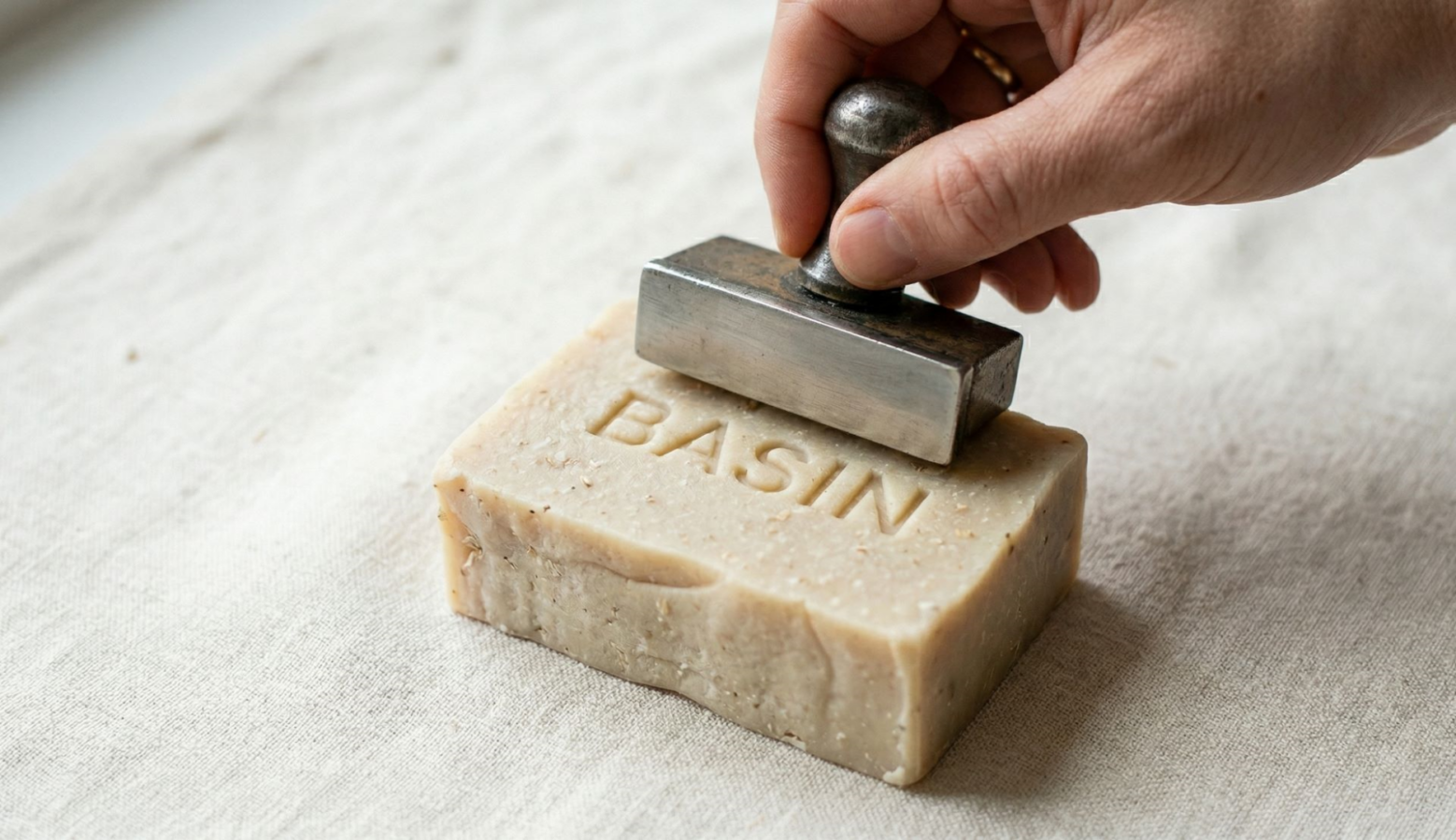 Close up of a hand pressing a metal stamp onto a beige soap bar with the word BASIN stamped on its surface