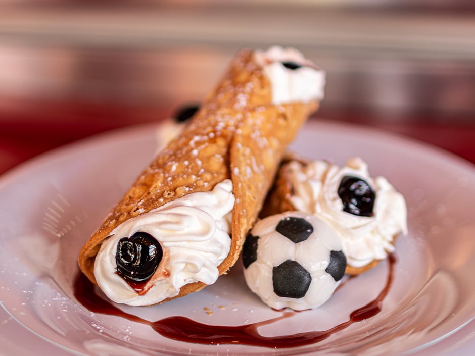 Two cannoli filled with whipped cream and cherry topping plated with a soccer ball shaped cookie and a sauce drizzle on a white plate