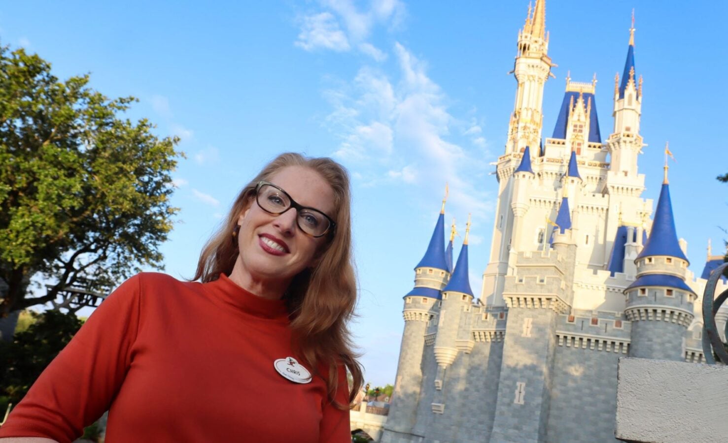 Smiling woman with glasses in a red top posing in front of a fairy tale castle with blue spires at a theme park