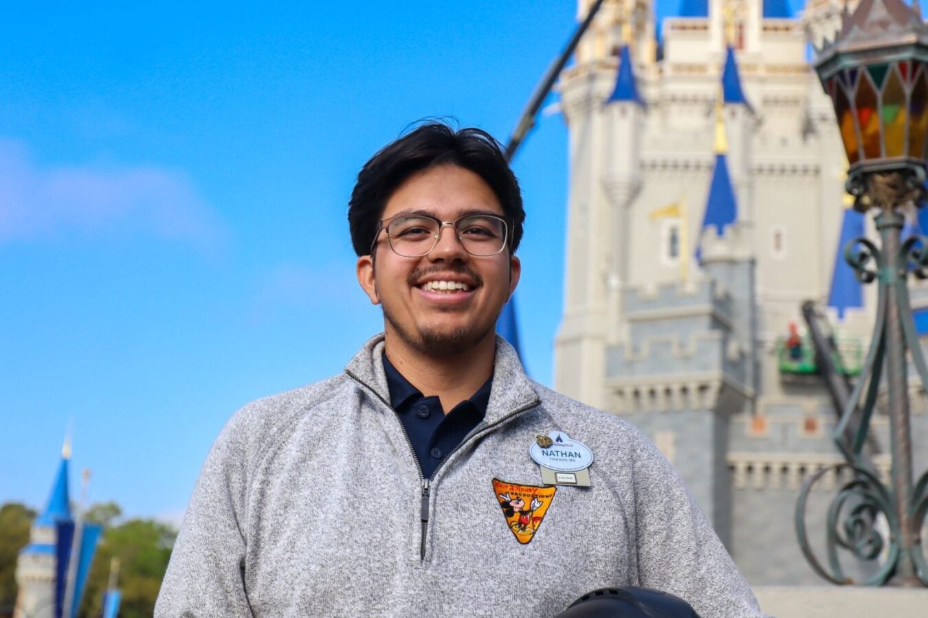 Smiling man with glasses in a gray zip up jacket wearing a name badge that says Nathan in front of a Disneyland castle