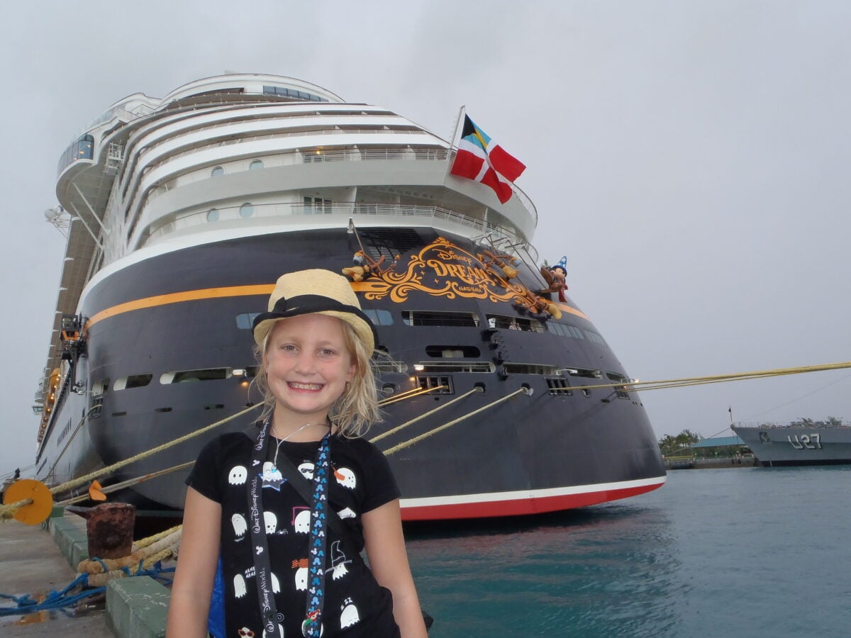 Smiling girl in front of a large Disney Dream cruise ship docked at the port with flags fluttering above the vessel