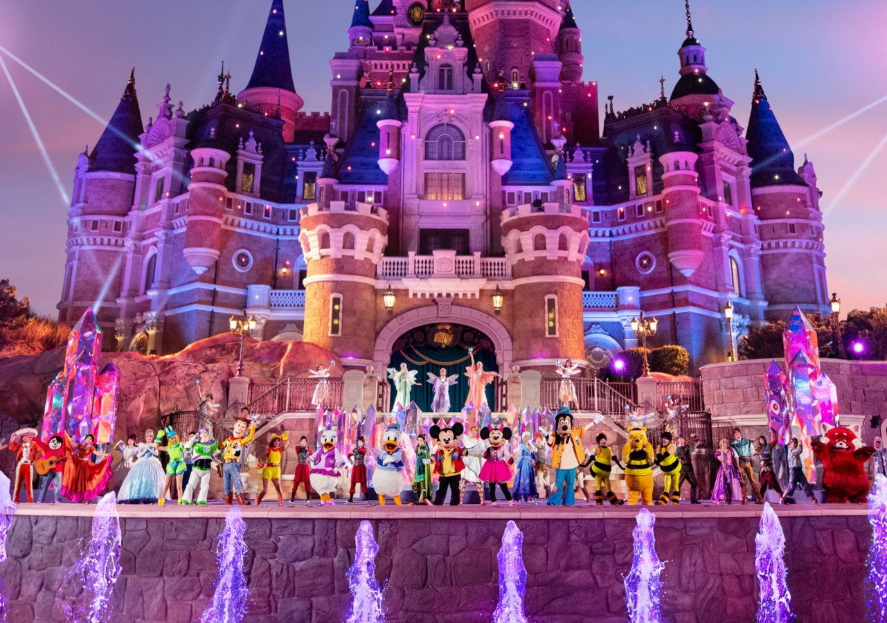 Castle lit in pink and blue with performers in colorful costumes on the stage and sparkling fountains in front at duskevening