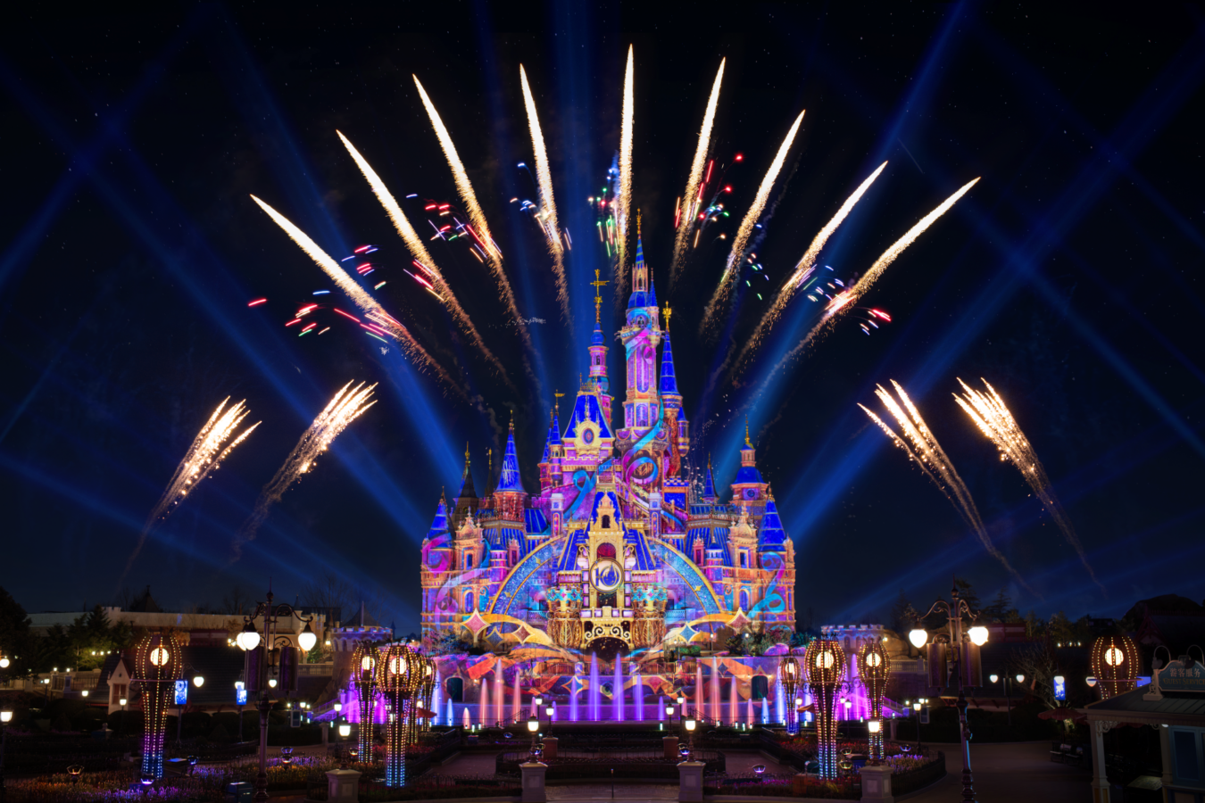 Castle illuminated with vibrant colorful lights at night as fireworks explode overhead behind it