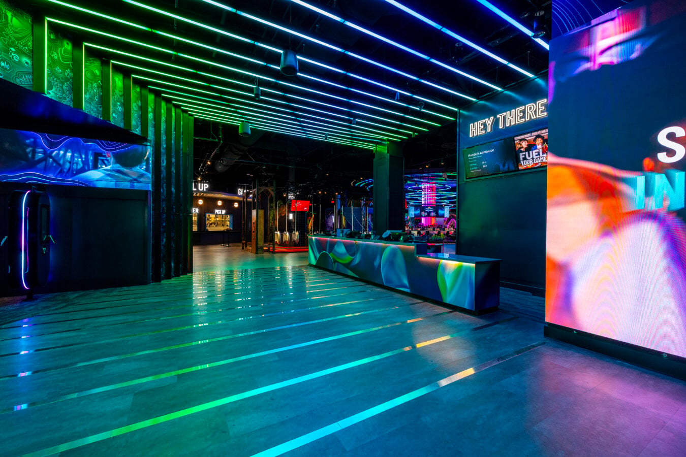 Interior of a neon lit arcade lobby with blue and green LED ceiling strips a colorful curved reception desk and a large screen reading HEY THERE