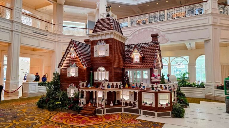 Grand hotel lobby with a tall gingerbread house centerpiece, decorated with windows, roof tiles, and holiday garlands at seating-height level among ornate railings.