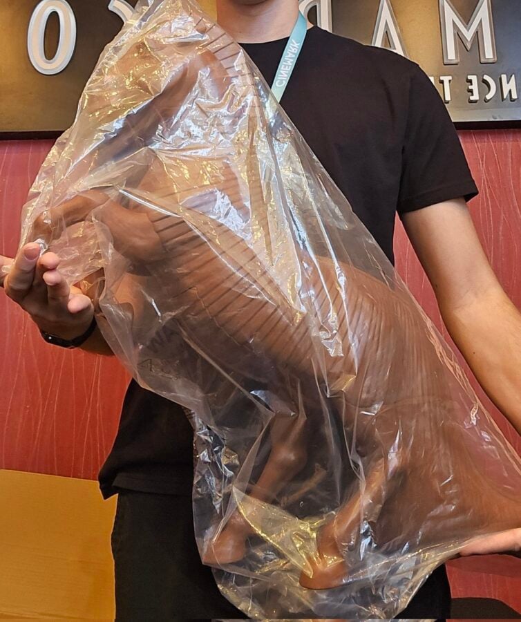 Person in a black shirt holding a large plastic wrapped piece of meat turkey leg toward the camera indoors wearing a light blue lanyard