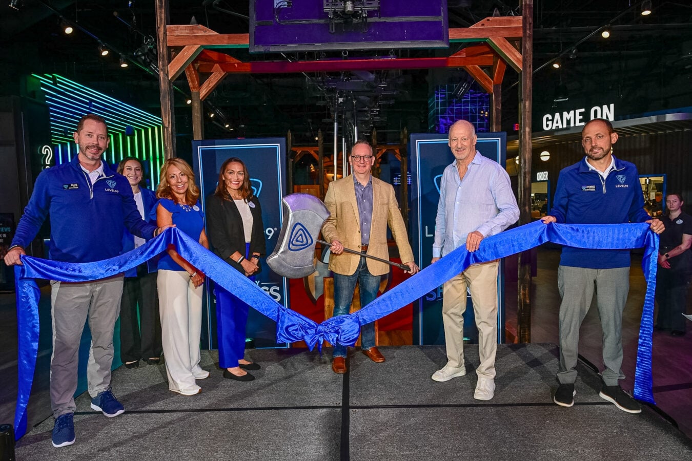 Seven people stand in a row at an indoor ribbon cutting ceremony holding a blue ribbon with a GAME ON sign visible in the background